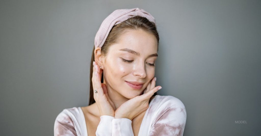 Beautiful woman smiles while holding her face and looking down. (Model)